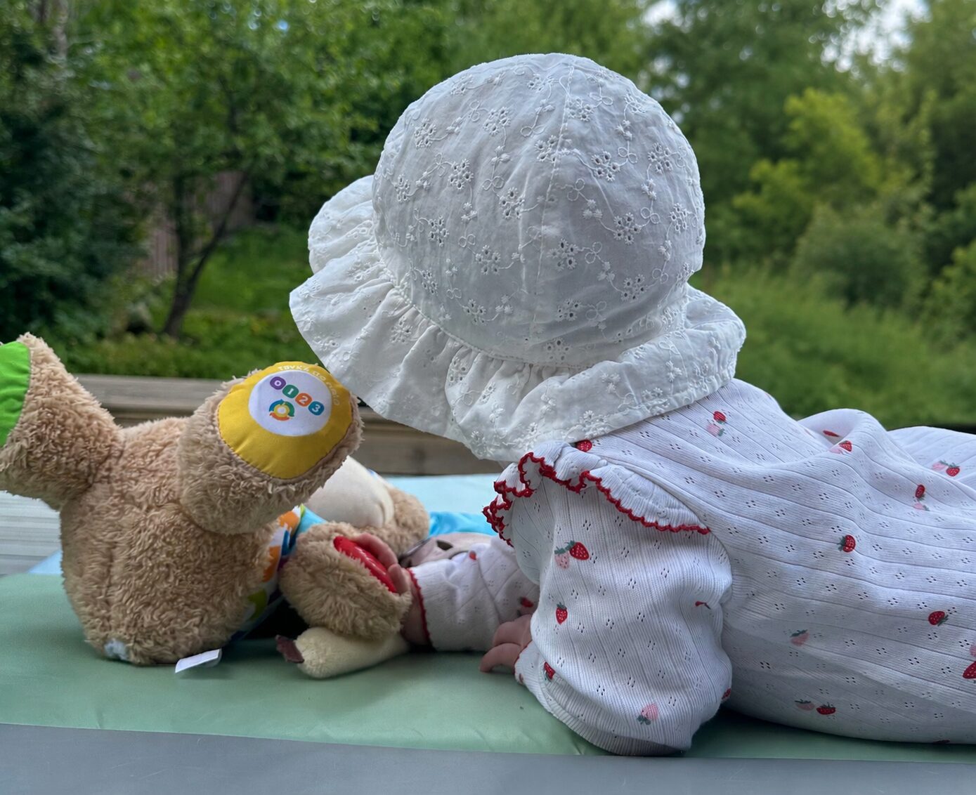 Baby i solhatt ligger på et skifteunderlag ute med Fisher-Price-valpen, sett bakfra